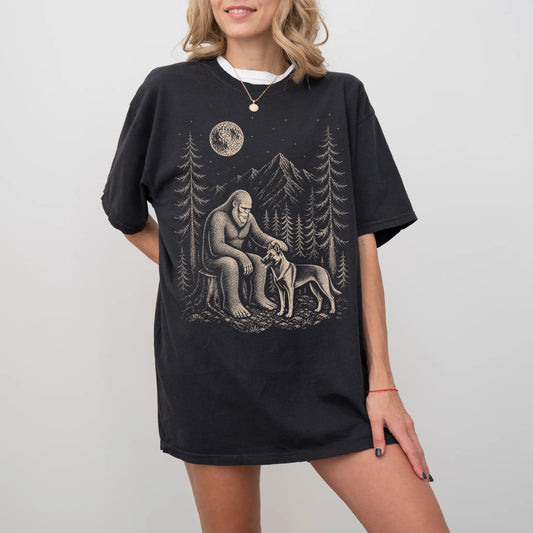 Model wearing a Black Comfort Colors® T-shirt with bigfoot petting a belgian malinois beneath the moon in a pine forest