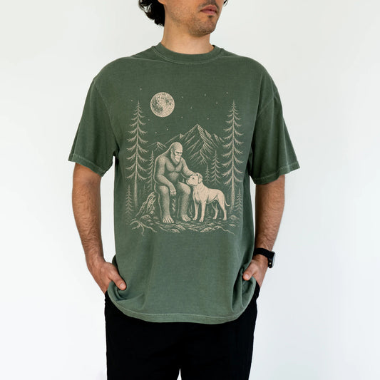 Model wearing a Moss Comfort Colors® T-shirt with bigfoot petting a american bulldog beneath the moon in a pine forest