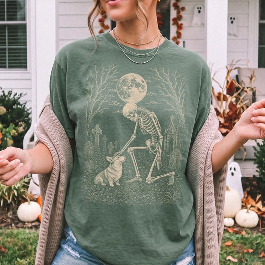 Model wearing a Moss Comfort Colors® T-shirt showing Corgi sharing a quiet cemetery moment with a friendly skeleton