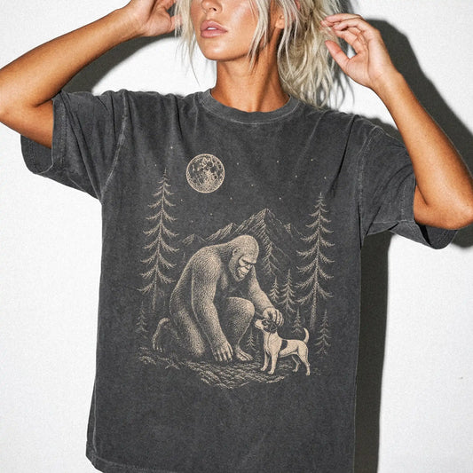 Model wearing a Pepper Comfort Colors® T-shirt with bigfoot petting a jack russell beneath the moon in a pine forest