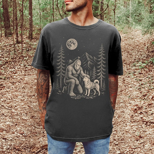 Model wearing a Pepper Comfort Colors® T-shirt with bigfoot petting a shiba inu beneath the moon in a pine forest