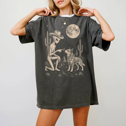 Model wearing a Pepper Comfort Colors® T-shirt with cowboy alien fist-bumping a blue heeler beneath the moon