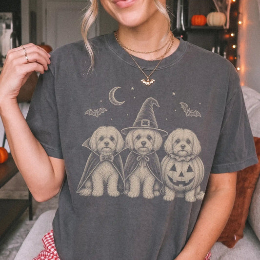 Model wearing a Pepper Comfort Colors® T-shirt showing three Maltese dogs dressed for Halloween beneath bats and a crescent m