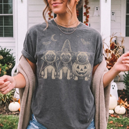 Model wearing a Pepper Comfort Colors® T-shirt showing three Pug dogs dressed for Halloween beneath bats and a crescent moon