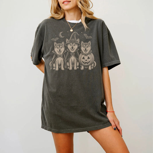 Model wearing a Pepper Comfort Colors® T-shirt showing three Shiba Inu dogs dressed for Halloween beneath bats and a crescent