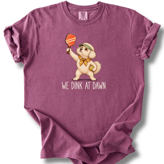 Bichon Frise with a pickleball paddle and ball on a Berry Comfort Colors® T-shirt with We Dink at Dawn text