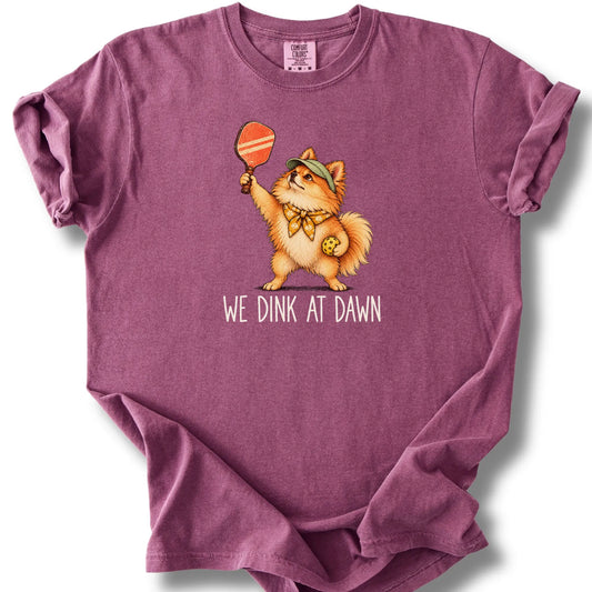 Pomeranian with a pickleball paddle and ball on a Berry Comfort Colors® T-shirt with We Dink at Dawn text