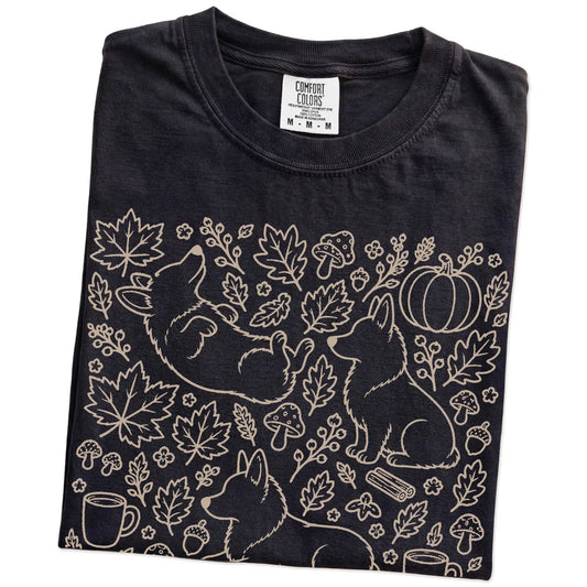 Folded Black Comfort Colors® T-shirt showing Corgi dogs with pumpkins, leaves, acorns, and woodland details