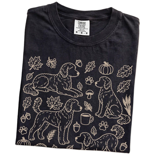 Folded Black Comfort Colors® T-shirt showing Goldendoodle dogs with pumpkins, leaves, acorns, and woodland details