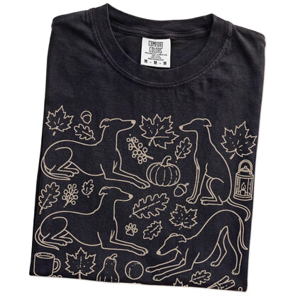 Folded Black Comfort Colors® T-shirt showing Greyhound dogs with pumpkins, leaves, acorns, and woodland details