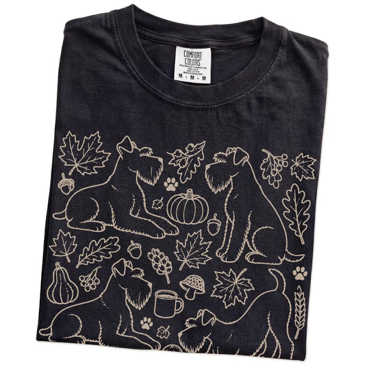 Folded Black Comfort Colors® T-shirt showing Schnauzer dogs with pumpkins, leaves, acorns, and woodland details