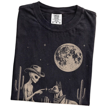 Folded Black Comfort Colors® T-shirt showing cowboy alien fist-bumping a belgian malinois beneath the moon