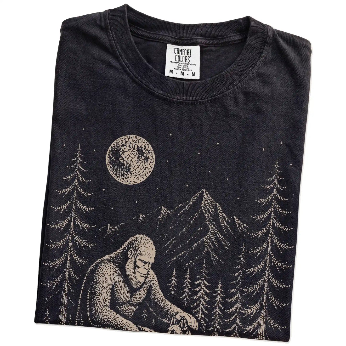 Folded Black Comfort Colors® T-shirt showing bigfoot petting a belgian malinois beneath the moon in a pine forest
