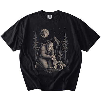 Bigfoot petting a Cavalier King Charles Spaniel beneath the moon in a pine forest on a Black Comfort Colors® T-shirt