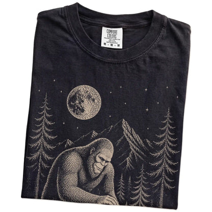Folded Black Comfort Colors® T-shirt showing bigfoot petting a cavalier king charles spaniel beneath the moon in a pine forest