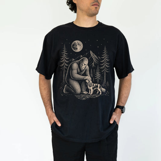 Model wearing a Black Comfort Colors® T-shirt with bigfoot petting a cavalier king charles spaniel beneath the moon in a pine forest