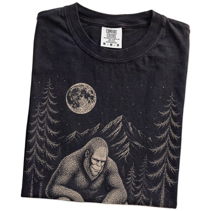 Folded Black Comfort Colors® T-shirt showing bigfoot petting a dachshund beneath the moon in a pine forest