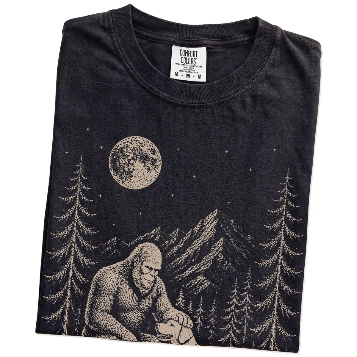 Folded Black Comfort Colors® T-shirt showing bigfoot petting a golden retriever beneath the moon in a pine forest