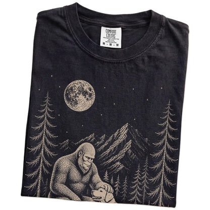 Folded Black Comfort Colors® T-shirt showing bigfoot petting a golden retriever beneath the moon in a pine forest
