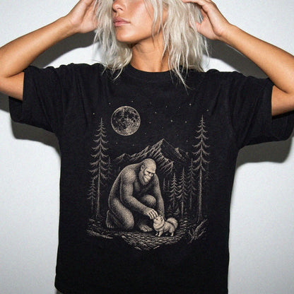 Model wearing a Black Comfort Colors® T-shirt with bigfoot petting a pomeranian beneath the moon in a pine forest