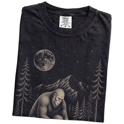 Folded Black Comfort Colors® T-shirt showing bigfoot petting a pomeranian beneath the moon in a pine forest