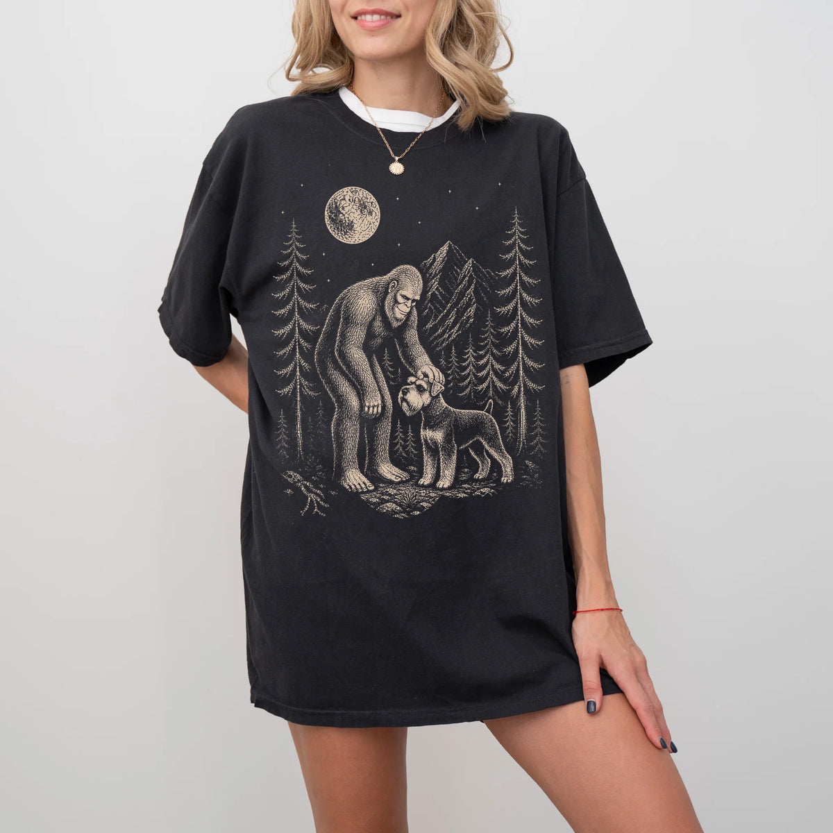 Model wearing a Black Comfort Colors® T-shirt with bigfoot petting a schnauzer beneath the moon in a pine forest