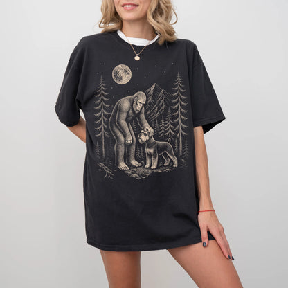 Model wearing a Black Comfort Colors® T-shirt with bigfoot petting a schnauzer beneath the moon in a pine forest