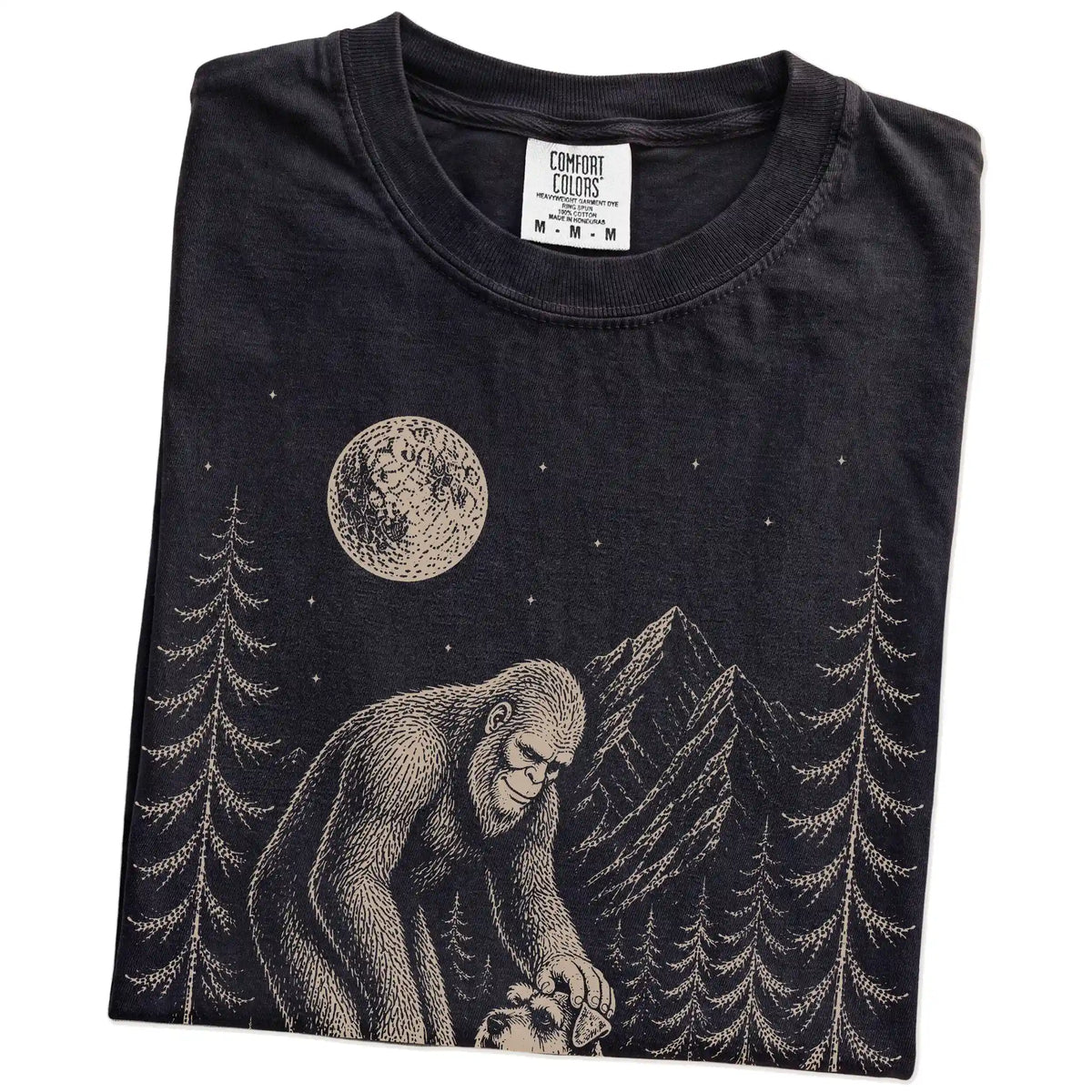 Folded Black Comfort Colors® T-shirt showing bigfoot petting a schnauzer beneath the moon in a pine forest
