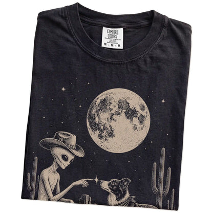 Folded Black Comfort Colors® T-shirt showing cowboy alien fist-bumping a border collie beneath the moon