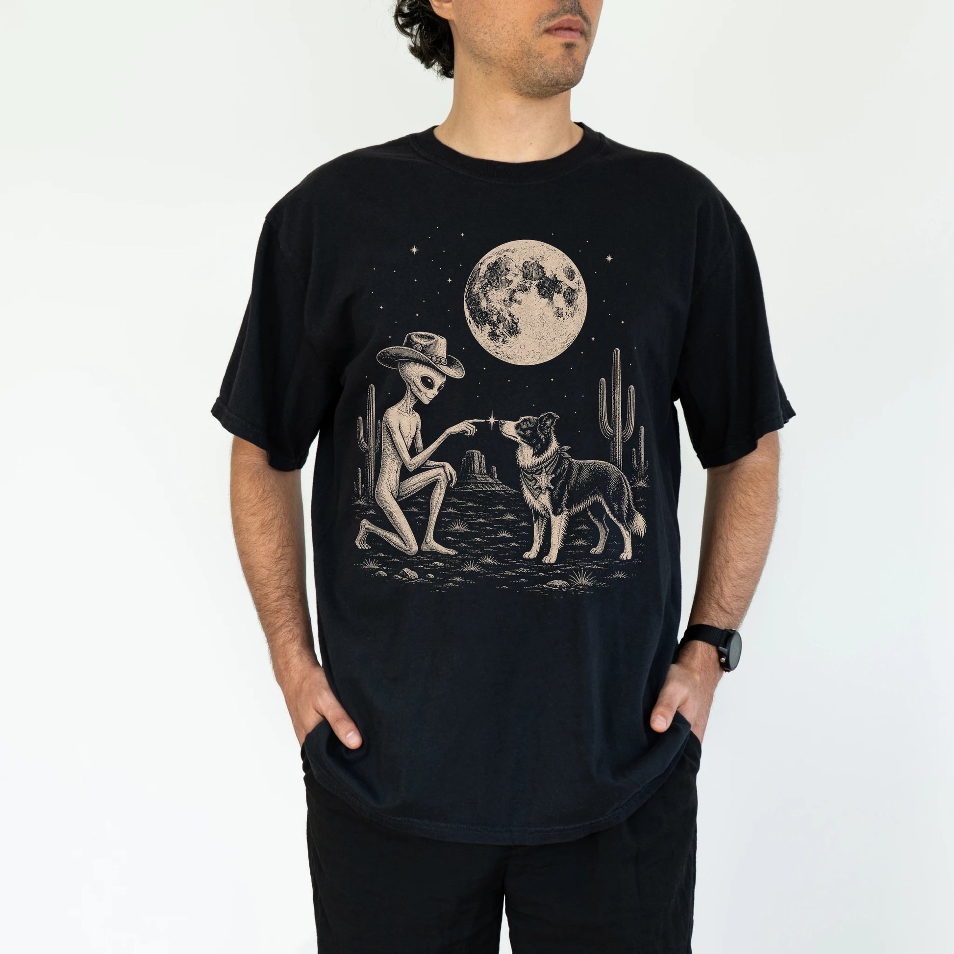 Model wearing a Black Comfort Colors® T-shirt with cowboy alien fist-bumping a border collie beneath the moon