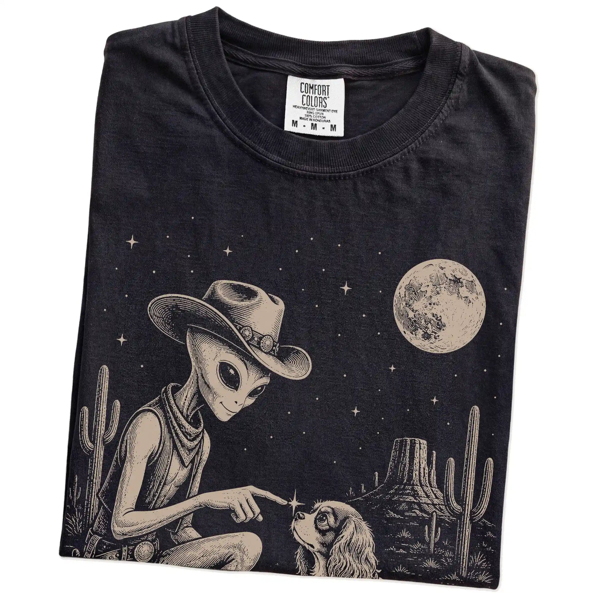 Folded Black Comfort Colors® T-shirt showing cowboy alien fist-bumping a cavalier king charles spaniel beneath the moon