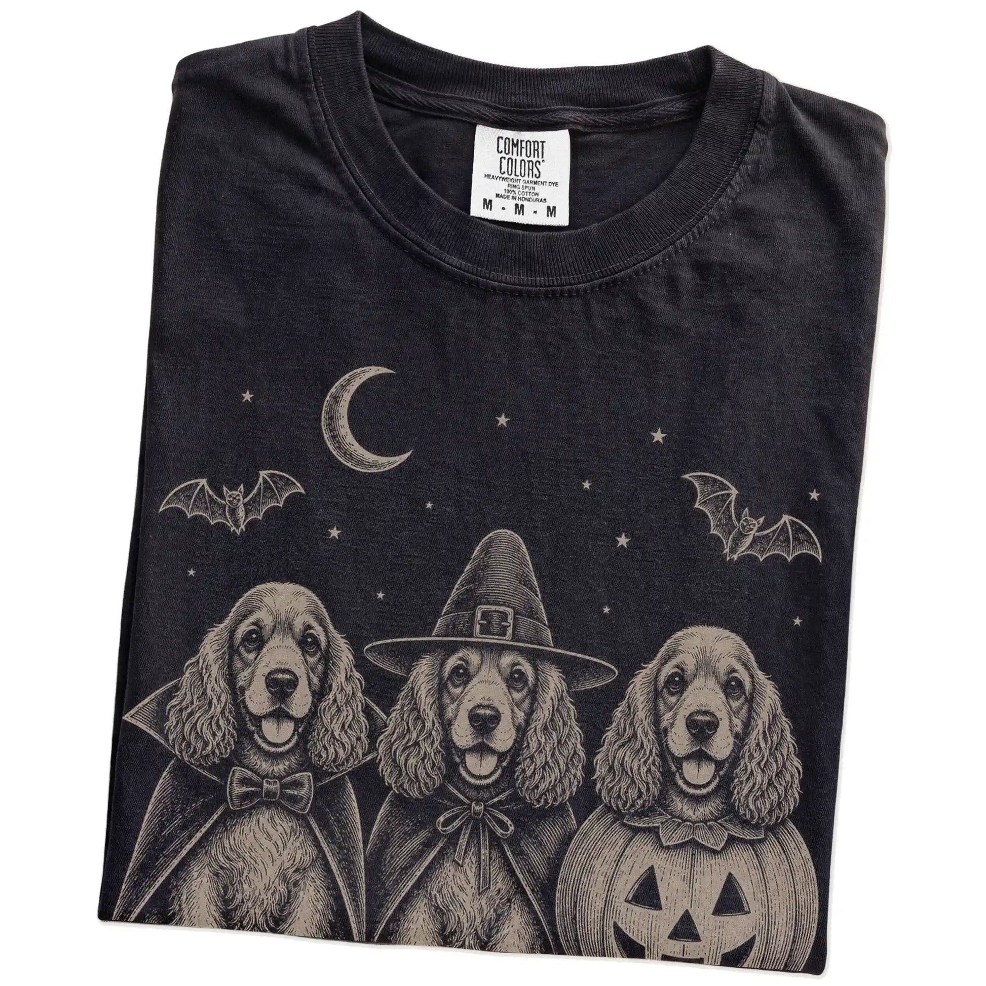 Folded Black Comfort Colors® T-shirt showing three Cocker Spaniel dogs dressed for Halloween beneath bats and a crescent moon