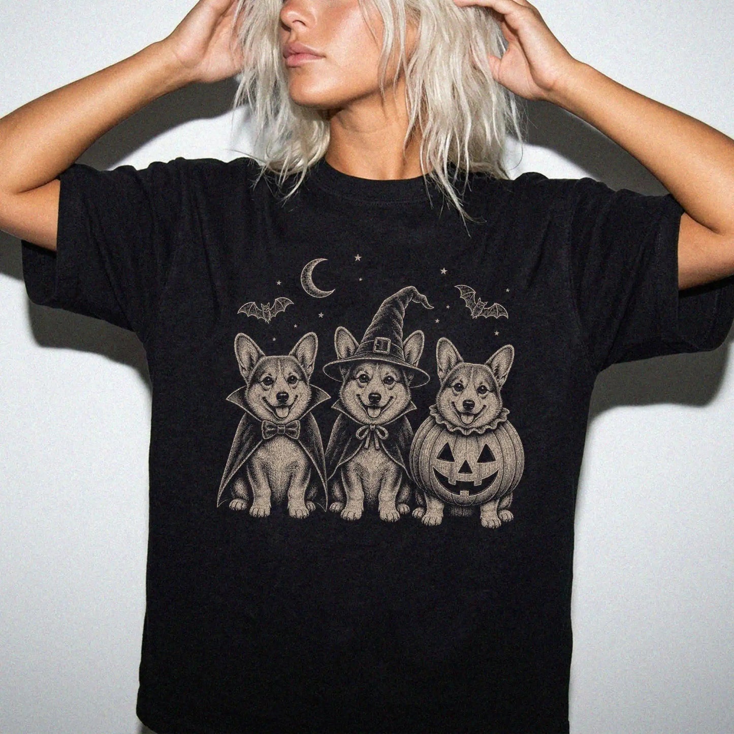 Model wearing a Black Comfort Colors® T-shirt showing three Corgi dogs dressed for Halloween beneath bats and a crescent moon
