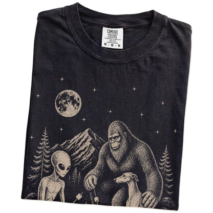 Folded Black Comfort Colors® T-shirt showing Greyhound camping with Bigfoot and an alien