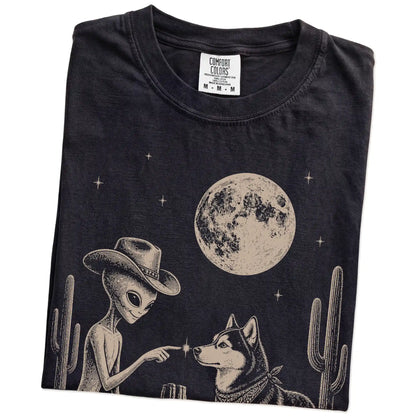 Folded Black Comfort Colors® T-shirt showing cowboy alien fist-bumping a husky beneath the moon