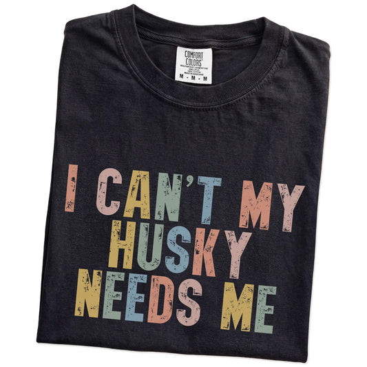 Folded Black Comfort Colors® T-shirt showing Husky with “I Can’t, My Dog Needs Me” lettering