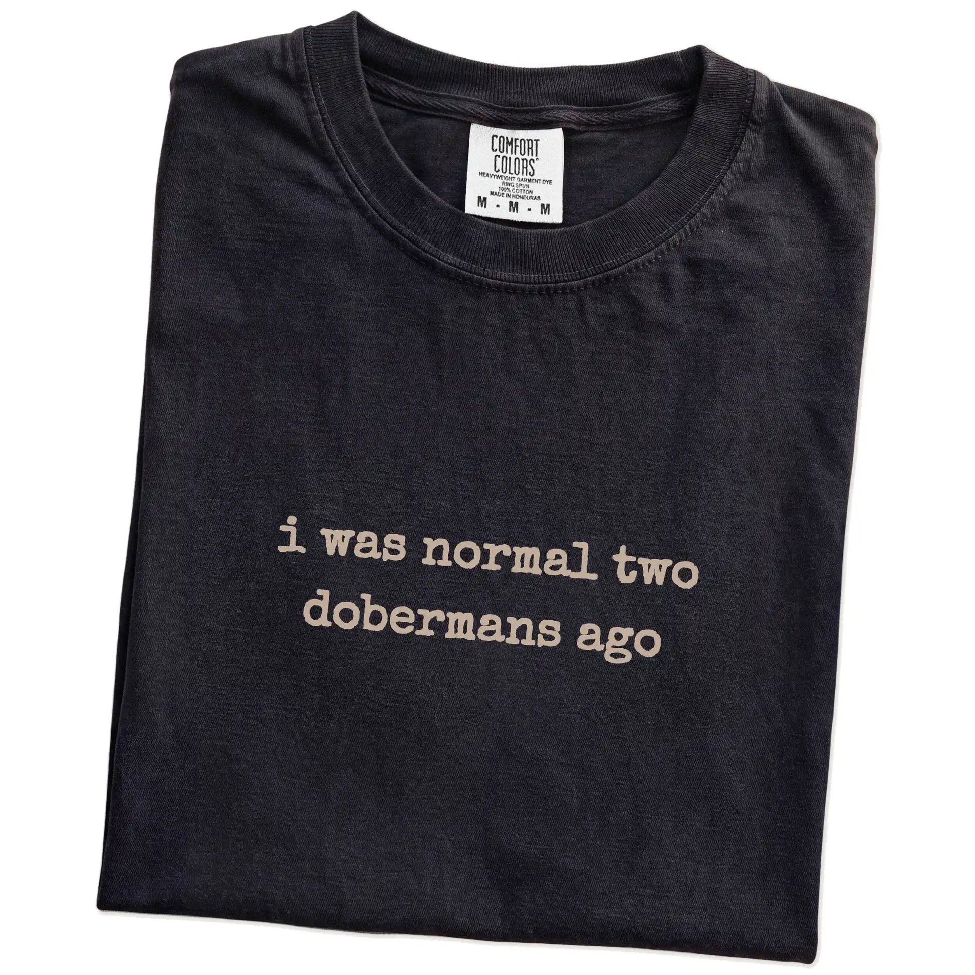 Folded Black Comfort Colors® T-shirt showing i was normal two dobermans ago lettering