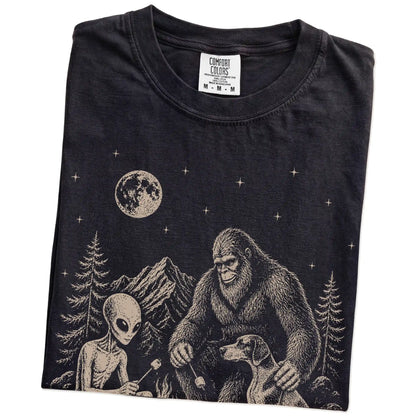Folded Black Comfort Colors® T-shirt showing Pointer camping with Bigfoot and an alien
