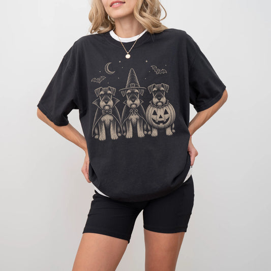 Model wearing a Black Comfort Colors® T-shirt showing three Schnauzer dogs dressed for Halloween beneath bats and a crescent 