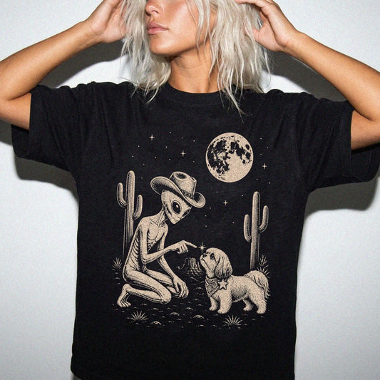 Model wearing a Black Comfort Colors® T-shirt with cowboy alien fist-bumping a shih tzu beneath the moon