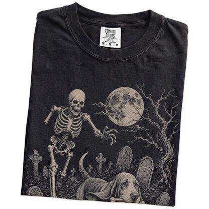 Folded Black Comfort Colors® T-shirt showing Basset Hound running through a moonlit graveyard with a chasing skeleton