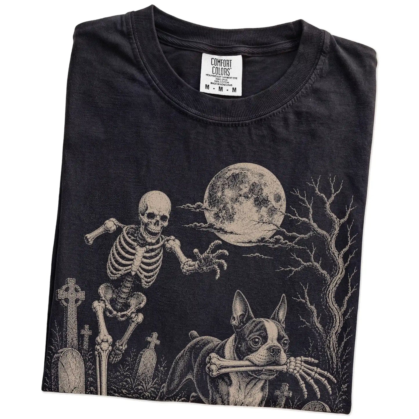 Folded Black Comfort Colors® T-shirt showing Boston Terrier running through a moonlit graveyard with a chasing skeleton