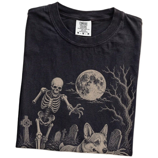 Folded Black Comfort Colors® T-shirt showing Corgi running through a moonlit graveyard with a chasing skeleton