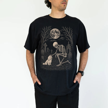 Model wearing a Black Comfort Colors® T-shirt showing Maltese sharing a quiet cemetery moment with a friendly skeleton