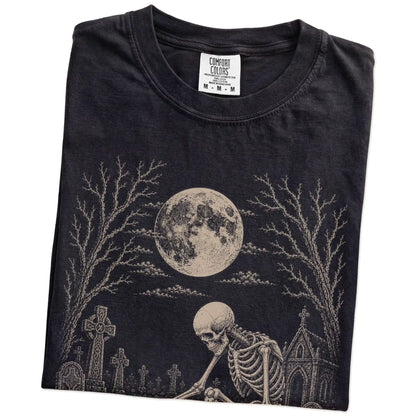 Folded Black Comfort Colors® T-shirt showing Pomeranian sharing a quiet cemetery moment with a friendly skeleton
