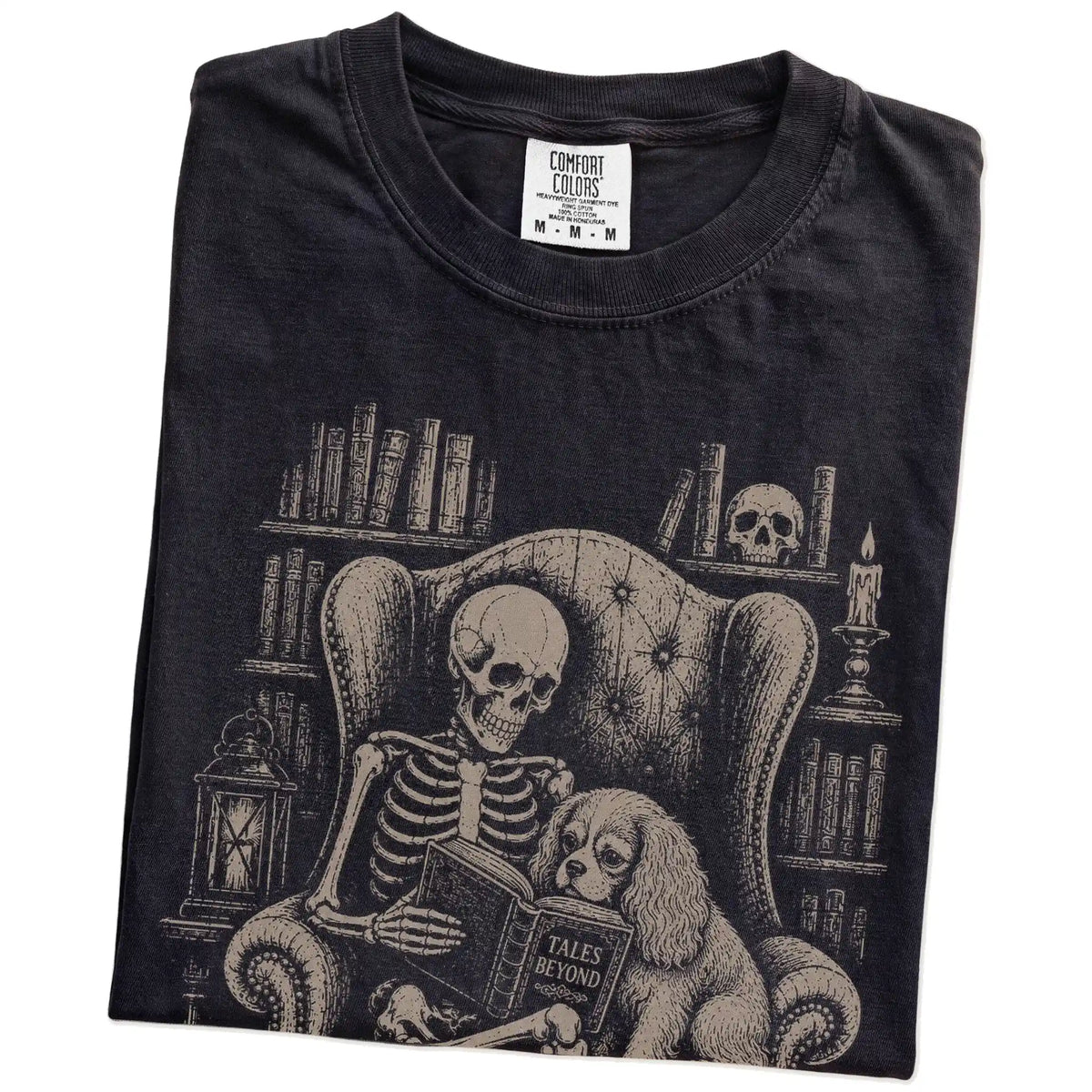 Folded Black Comfort Colors® T-shirt showing skeleton reading in a library beside a Cavalier King Charles Spaniel