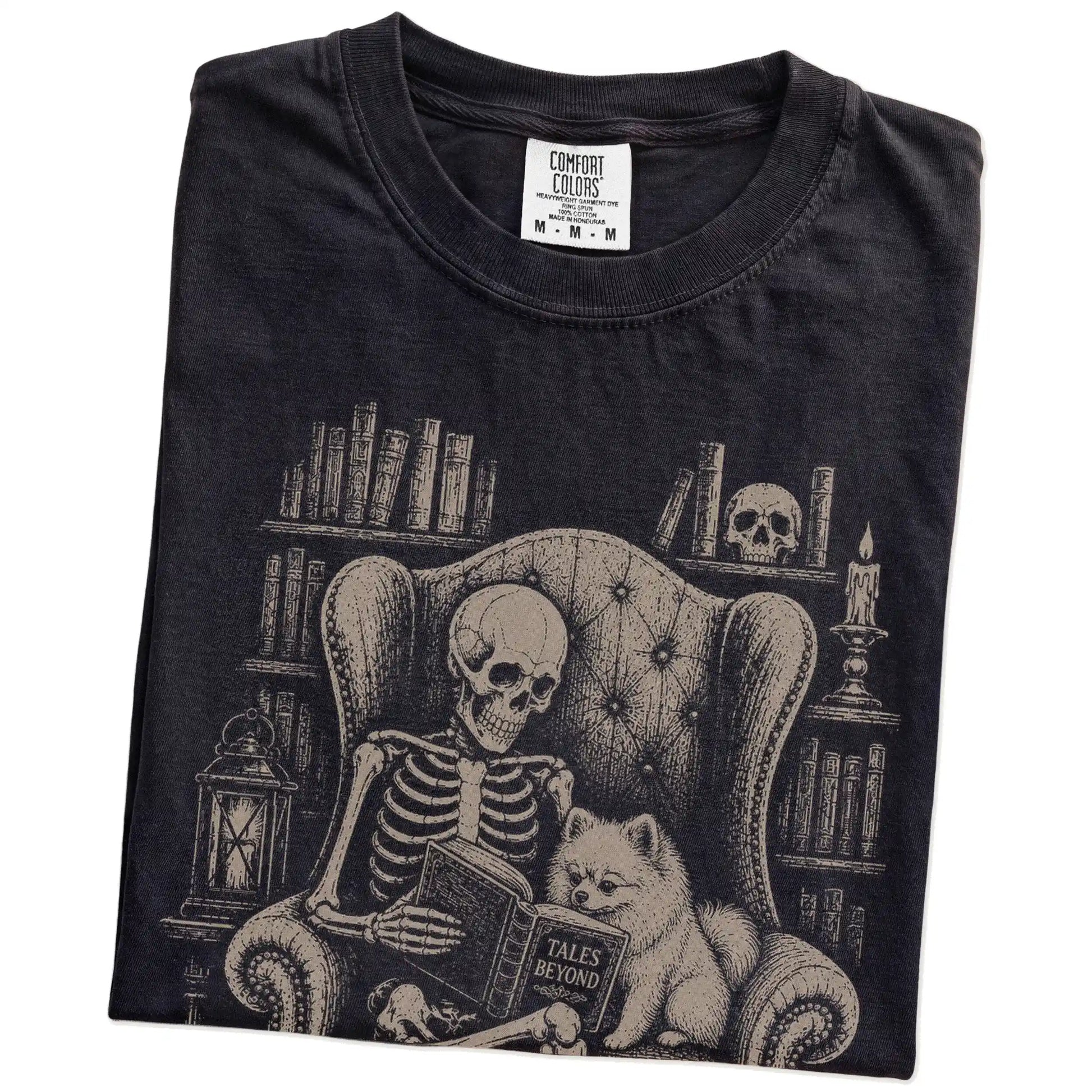 Folded Black Comfort Colors® T-shirt showing skeleton reading in a library beside a Pomeranian