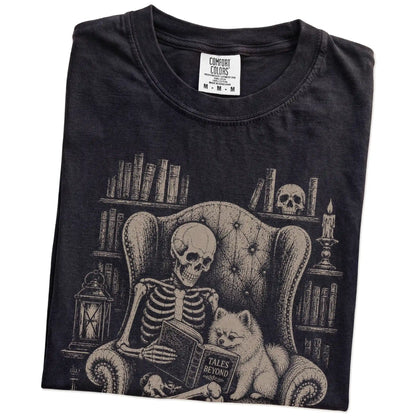 Folded Black Comfort Colors® T-shirt showing skeleton reading in a library beside a Pomeranian