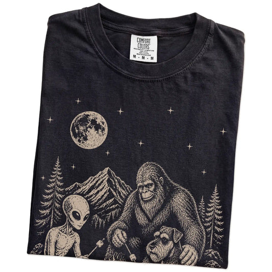 Folded Black Comfort Colors® T-shirt showing Standard Schnauzer camping with Bigfoot and an alien