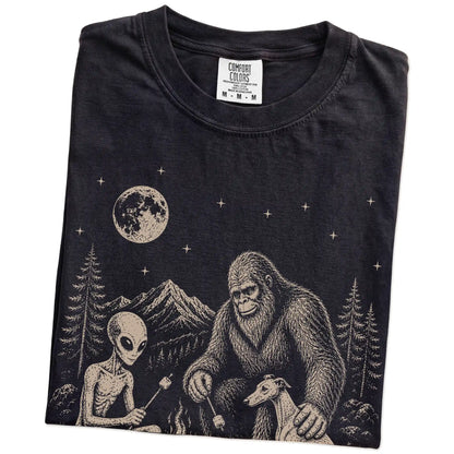 Folded Black Comfort Colors® T-shirt showing Whippet camping with Bigfoot and an alien